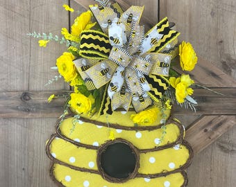 May include: A decorative, yellow beehive-shaped wreath with a polka-dot pattern. The wreath is adorned with yellow flowers and a large bow featuring bee and honeycomb designs. The wreath is approximately 15 inches in diameter.