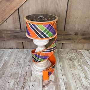 May include: A spool of decorative ribbon with a plaid pattern in orange, purple, green, and black. The ribbon is displayed on a white pedestal. The ribbon is unrolled, showing the pattern and texture.