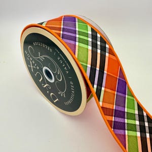 May include: A spool of D'Lux ribbon with a plaid pattern in orange, purple, green, black, and white. The ribbon has an orange border and is labeled "D'Lux Ribbon" with the words "Value" and "Sophisticated".