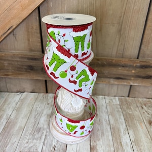 May include: A roll of white wired ribbon featuring a repeating pattern of green furry legs, red Santa hats, and colorful Christmas ornaments. The ribbon edges are finished with a shimmering red glitter trim, creating a festive holiday craft supply.