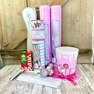 May include: A craft supply set featuring two rolls of pink iridescent foil mesh, a roll of thin stripe fabric mesh, three spools of holiday ribbon, a box of 100 mini lights, white chenille stems, pink ornaments, and a pink top hat decoration.