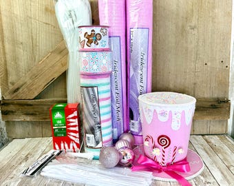 May include: A craft supply set featuring two rolls of pink iridescent foil mesh, a roll of thin stripe fabric mesh, three spools of holiday ribbon, a box of 100 mini lights, white chenille stems, pink ornaments, and a pink top hat decoration.