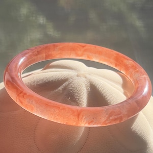 May include: A smooth, circular bangle bracelet featuring a translucent, marbled appearance with swirling shades of coral, peach, and soft orange. The material has a polished, glossy finish that catches the light, creating a semi-transparent effect.