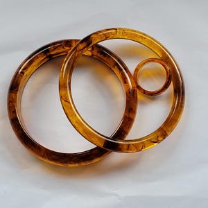 May include: Three amber-colored, circular bangles. The largest bangle is approximately 4 inches in diameter, the second is about 3 inches, and the smallest is 1 inch. The bangles have a marbled, tortoiseshell-like pattern.