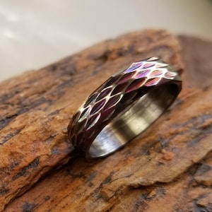 May include: A dark grey metal ring with a textured, geometric pattern. The ring has a brushed finish and a subtle iridescent sheen.