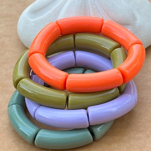 May include: Four colorful bracelets in shades of orange, olive green, lavender, and teal. Each bracelet is made of rounded, cylindrical beads. A white, textured object is in the background.
