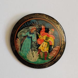 May include: A round black lacquered wooden pin with a gold trim. The pin features a painted scene of a woman in a blue dress and a man in a red and yellow outfit. The scene is set against a dark blue background with white and gold accents. The pin is approximately 2 inches in diameter.