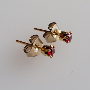 May include: A pair of gold-colored stud earrings, each featuring a small, round, deep red gemstone. The earrings have a classic post-and-clutch design, with a simple, elegant aesthetic. The gemstones are securely set in a gold-colored metal.