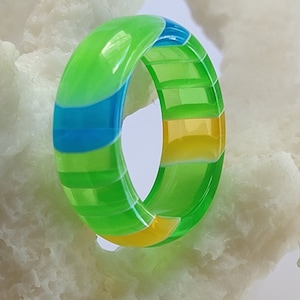 May include: A green, yellow, and blue striped ring made of resin. The ring is glossy and has a smooth finish.