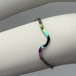May include: A minimalist bracelet with a wavy design. The bracelet has a metallic finish with a rainbow color gradient, transitioning from purple to green. The bracelet is worn on a white arm.