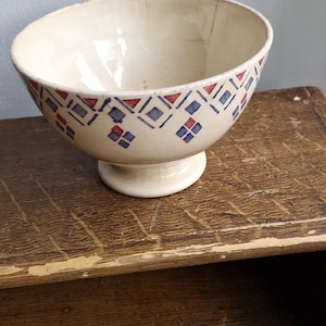 May include: Cream-coloured ceramic bowl with a pedestal base. The bowl has a geometric pattern of red and blue diamonds and triangles around the rim. The bowl is displayed on a wooden surface.