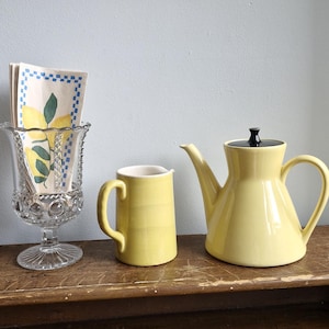 May include: A still life arrangement featuring a yellow teapot with a black lid, a matching creamer, and a decorative glass holder with lemon-themed napkins. A framed abstract artwork hangs in the background. The objects are on a wooden surface.