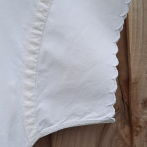 May include: Close-up of a white garment sleeve with a scalloped edge. The sleeve is made of a soft, textured fabric. The edge of the sleeve is scalloped. The background is a wooden fence.