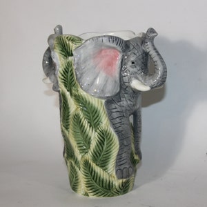 May include: A ceramic vase shaped like an elephant with green leaves. The elephant is gray with a pink ear and a white tusk.