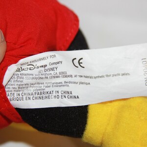 Puede incluir: Una etiqueta blanca con texto negro unida a un juguete de peluche rojo, negro y amarillo. La etiqueta dice "Fabricado exclusivamente para Walt Disney Company &copy; Disney" e incluye informaci&oacute;n sobre los materiales utilizados y el pa&iacute;s de origen. La etiqueta tambi&eacute;n incluye la marca CE y el n&uacute;mero "DN 20401".