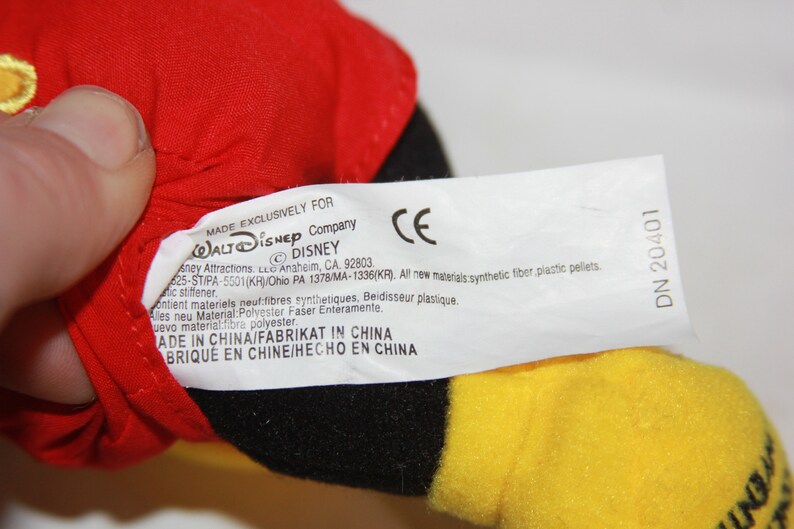 Puede incluir: Una etiqueta blanca con texto negro unida a un juguete de peluche rojo, negro y amarillo. La etiqueta dice "Fabricado exclusivamente para Walt Disney Company &copy; Disney" e incluye informaci&oacute;n sobre los materiales utilizados y el pa&iacute;s de origen. La etiqueta tambi&eacute;n incluye la marca CE y el n&uacute;mero "DN 20401".