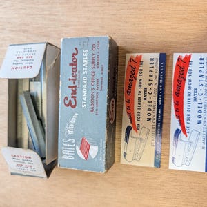May include: An assortment of vintage office supplies, including a box of "End-icator" standard staples, a box of staples, and two promotional cards for a "Model C Stapler." The boxes are light blue and white.