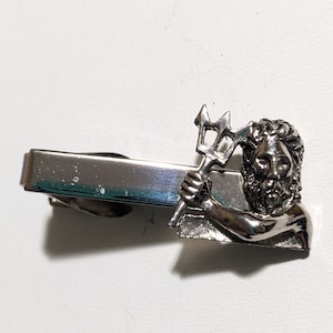 May include: Silver tie clip featuring a detailed depiction of a mythical figure holding a trident.
