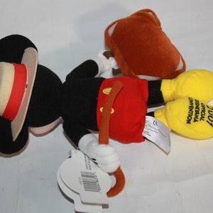 Puede incluir: Un juguete de peluche de Mickey Mouse con un sombrero de paja y pantalones cortos rojos. El juguete sostiene un palo marr&oacute;n y tiene una maleta amarilla. El juguete est&aacute; etiquetado como "2007 Convenci&oacute;n Oficial de Disneyland"