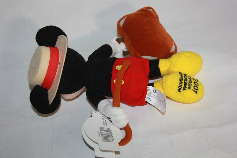 Puede incluir: Un juguete de peluche de Mickey Mouse con un sombrero de paja y pantalones cortos rojos. El juguete sostiene un palo marr&oacute;n y tiene una maleta amarilla. El juguete est&aacute; etiquetado como "2007 Convenci&oacute;n Oficial de Disneyland"