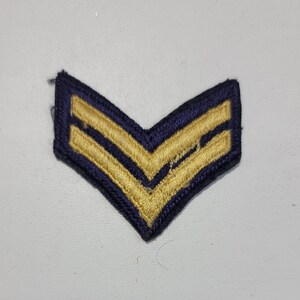May include: A navy blue chevron patch with two gold stripes. The patch is made of fabric and has a stitched edge.