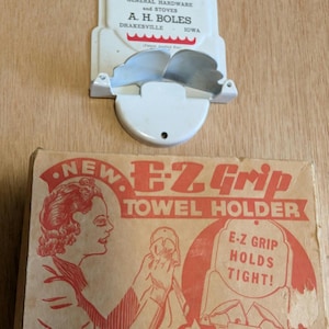May include: Vintage towel holder and hardware store sign. The towel holder is white with a metal grip. The sign is white with red text that reads "We Hold To The Best GENERAL HARDWARE and STOVES A.H. BOLES DRAKESVILLE IOWA". The towel holder box reads "E-Z Grip TOWEL HOLDER".