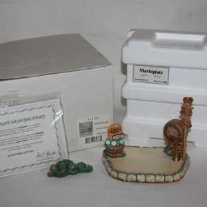 May include: A white box with a certificate of authenticity for a Marketplace figurine. The figurine is a small, detailed scene of a marketplace with a basket of apples, a barrel, and a sign that says "Apples". The figurine is made of ceramic and is painted in realistic colors. The box is also white and has a label that says "Marketplace".