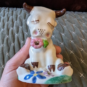 May include: A ceramic cow figurine with closed eyes, brown horns, and a pink flower collar. The white cow has brown hooves and is sitting on a green base with blue and purple flowers. A charming, vintage-style decorative piece.