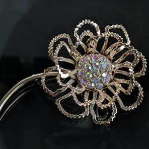 May include: A gold-toned flower brooch with a central cluster of iridescent rhinestones. The brooch features a detailed, openwork design with scalloped edges, and a curved pin for attachment. The flower's petals are intricately shaped.
