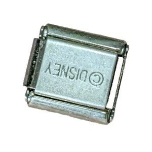 Puede incluir: Un charm de metal plateado con la palabra "DISNEY" y un s&iacute;mbolo de copyright.