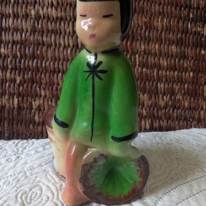 May include: A ceramic figurine of a seated figure in a green coat with black trim and a black hairstyle. The figurine has a light skin tone and is holding a basket. The figurine is sitting on a green leaf.