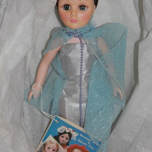 May include: A porcelain doll wearing a silver dress and a blue tulle cape. The doll has brown hair, green eyes, and a crown on her head. The doll is holding a small white bag in her hand. The doll is sitting on a white background. The doll is part of a collection of "Wonderful World of Effanbee Dolls".