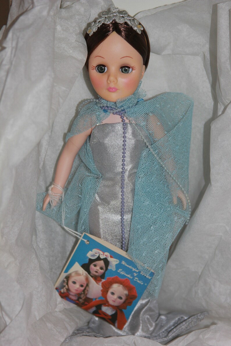 May include: A porcelain doll wearing a silver dress and a blue tulle cape. The doll has brown hair, green eyes, and a crown on her head. The doll is holding a small white bag in her hand. The doll is sitting on a white background. The doll is part of a collection of "Wonderful World of Effanbee Dolls".