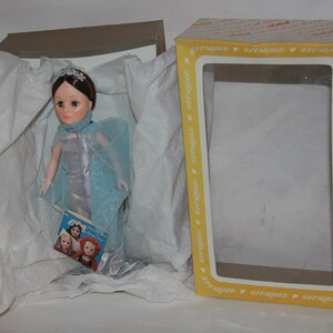 May include: A porcelain doll wearing a blue and silver gown with a matching cape. The doll is in a white tissue-filled box with a yellow and white box lid. The doll is holding a book with a picture of three dolls on the cover.