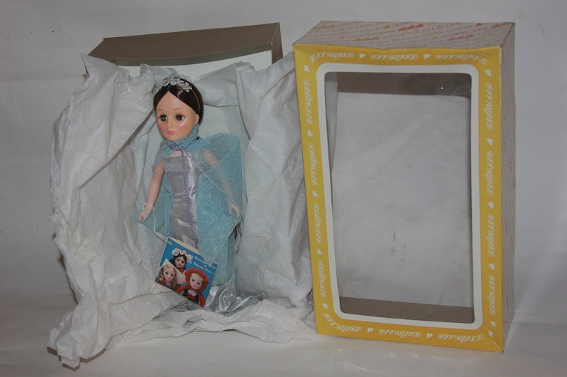 May include: A porcelain doll wearing a blue and silver gown with a matching cape. The doll is in a white tissue-filled box with a yellow and white box lid. The doll is holding a book with a picture of three dolls on the cover.