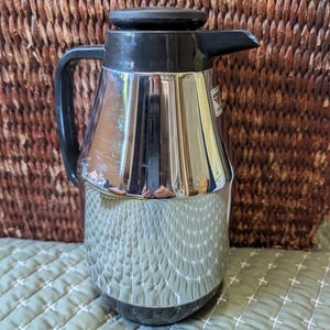 May include: A vintage chrome and black insulated carafe with a handle and spout. The carafe has a ribbed design on the lower half and a black lid. The carafe is likely designed to keep liquids hot or cold.