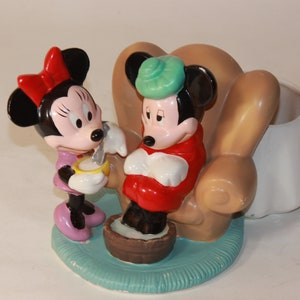 May include: A ceramic figurine set featuring Mickey Mouse and Minnie Mouse. Mickey is sitting in a brown armchair with a green ice pack on his head. Minnie is standing next to him, holding a spoon and a bowl. The set is a whimsical depiction of Mickey getting sick and Minnie taking care of him.