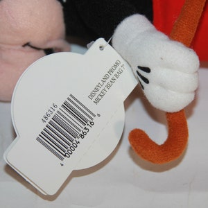 Puede incluir: Una etiqueta blanca con texto negro y un c&oacute;digo de barras que dice "DISNEYLAND PROMO MICKEY BEAN BAG '77" y "486316 00004 86316 6". La etiqueta est&aacute; unida a un juguete de peluche de Mickey Mouse con una camisa roja y un palo marr&oacute;n.