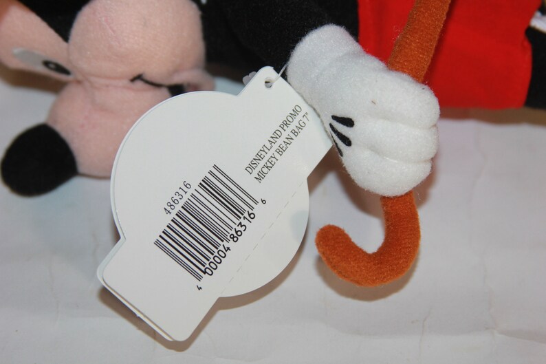 Puede incluir: Una etiqueta blanca con texto negro y un c&oacute;digo de barras que dice "DISNEYLAND PROMO MICKEY BEAN BAG '77" y "486316 00004 86316 6". La etiqueta est&aacute; unida a un juguete de peluche de Mickey Mouse con una camisa roja y un palo marr&oacute;n.