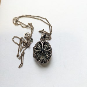 May include: A silver locket necklace with a detailed floral design and black accents. The locket is oval shaped and hangs from a silver chain.