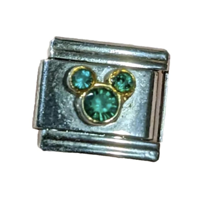 Puede incluir: Un charm para pulsera de plata con un dise&ntilde;o de cabeza de Mickey Mouse en oro con tres diamantes de imitaci&oacute;n verdes.