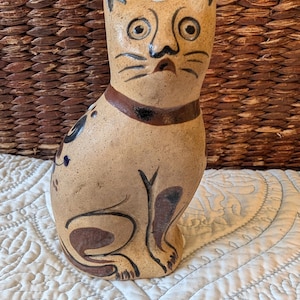 May include: A tan ceramic cat figurine with black and brown painted details. The cat has large eyes, a brown collar, and painted whiskers. The figurine is sitting upright and has a whimsical, folk art style.