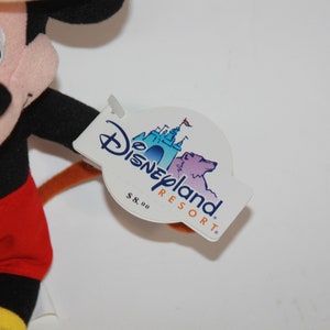 Puede incluir: Un juguete de peluche de Mickey Mouse con pantalones cortos rojos y zapatos amarillos. El juguete tiene una cola marr&oacute;n y una etiqueta que dice "Disneyland Resort" con un precio de 8,00&nbsp;&euro;.