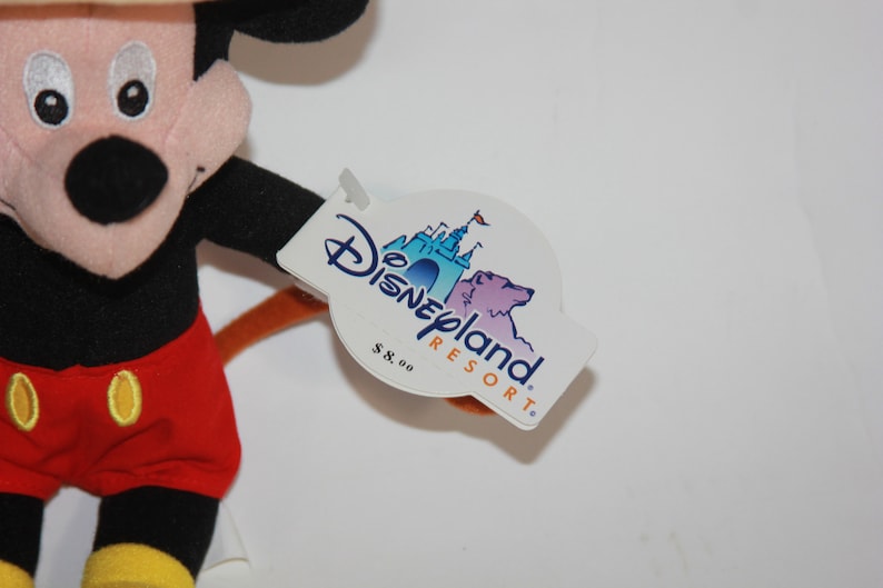 Puede incluir: Un juguete de peluche de Mickey Mouse con pantalones cortos rojos y zapatos amarillos. El juguete tiene una cola marr&oacute;n y una etiqueta que dice "Disneyland Resort" con un precio de 8,00&nbsp;&euro;.