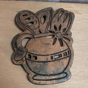 May include: A copper-colored metal trivet in the shape of a vase with flowers. The trivet has a handle and decorative cutouts. The vase has a horizontal band of rectangular cutouts. The flowers include a daisy and leaves.