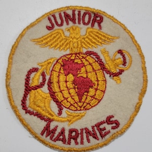 May include: A white circular patch with the words "Junior Marines" in red lettering around the edge. A gold eagle with outstretched wings is above a globe with red continents and a gold anchor.
