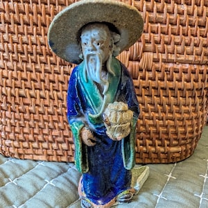 May include: A ceramic figurine of an elderly Asian man wearing a wide-brimmed hat and a long robe. The robe is primarily blue with green and gold accents. The figure is holding a textured object, possibly a basket or container.