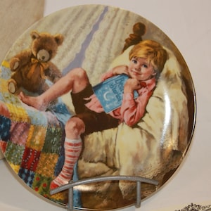 May include: A porcelain plate with a colorful illustration of a boy in bed reading a book with a teddy bear beside him. The book is titled "Bedtime Stories".