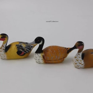 May include: Three wooden duck figurines painted in yellow, orange, and brown with black and white accents. The ducks are arranged in a row on a white surface.