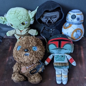 May include: A collection of Star Wars plush toys featuring characters like Yoda, Kylo Ren, BB-8, Chewbacca, and Boba Fett. The plushies are in various colors, including green, brown, black, white, and orange, and are arranged on a dark wooden surface.